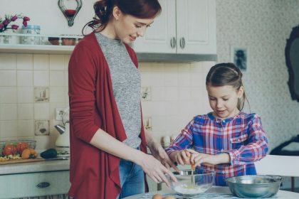 Little cheerful girl helping her mother in the kitchen cropping egg into bowl and mixing dough for cookies. Family, food, home and people concept