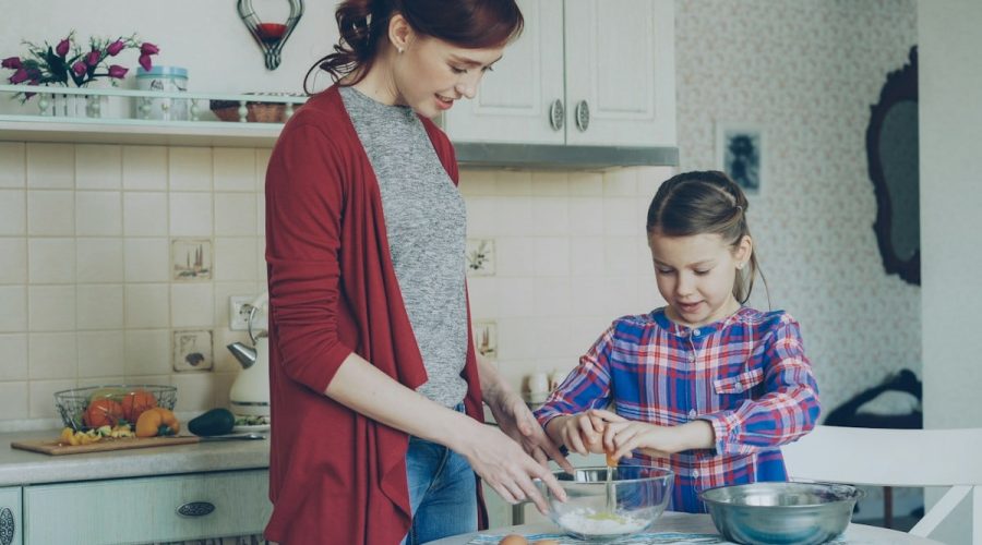 Little cheerful girl helping her mother in the kitchen cropping egg into bowl and mixing dough for cookies. Family, food, home and people concept