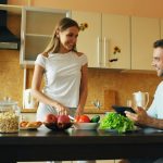 Attractive couple chatting in the kitchen early morning. Handsome man using tablet while his girlfriend cooking breakfast at home